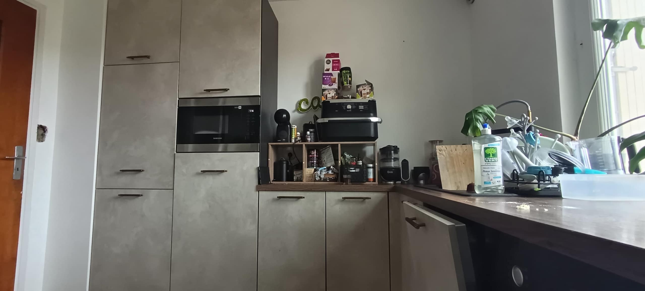 Modern kitchen with tall grey cabinets and a built-in microwave; a printer sits on a wooden shelf amid jars and small appliances.