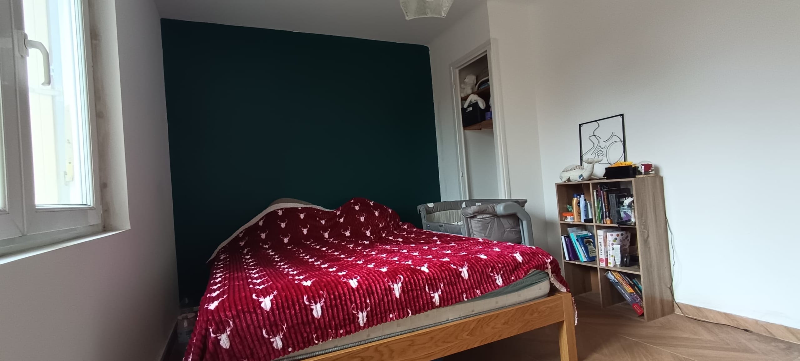 Bedroom with a wooden bed and red patterned blanket, teal accent wall, window on the left, and a bookshelf on the right edge of the room.