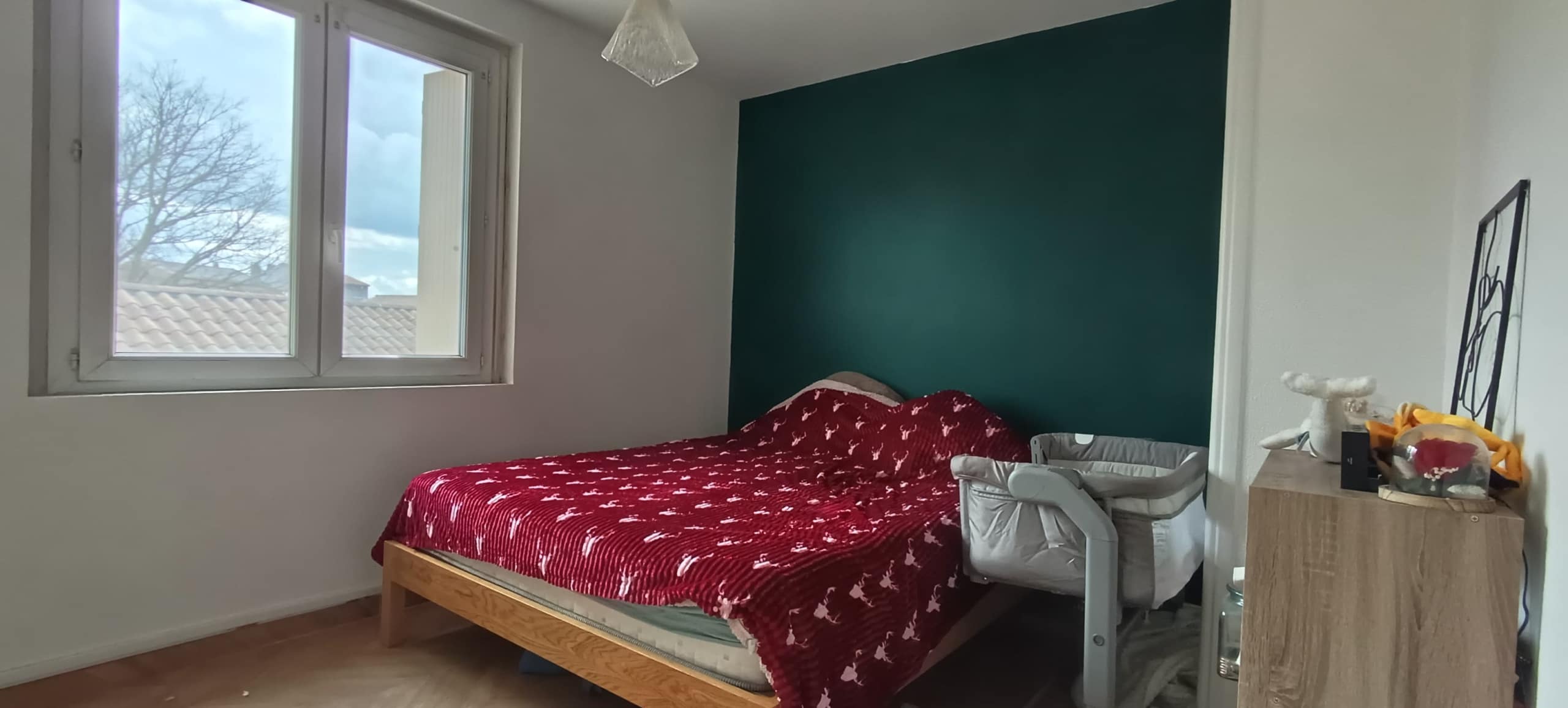 Single bed with a red patterned quilt against a teal accent wall, a window to the left letting in light, and a dresser with objects on top to the right.