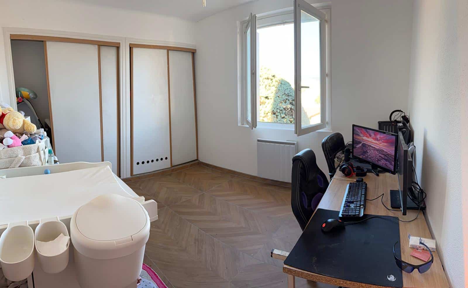 Bedroom with a computer desk setup on the right: monitor, keyboard, mouse, and headphones; open window letting in daylight; wooden parquet floor and large sliding wardrobes on the left; a storage bin with stuffed toys near a white changing table-like surface.