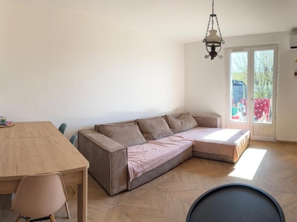 Cozy living room with a beige L-shaped sofa, a long wooden dining table, chairs, and a balcony door letting sunlight in.