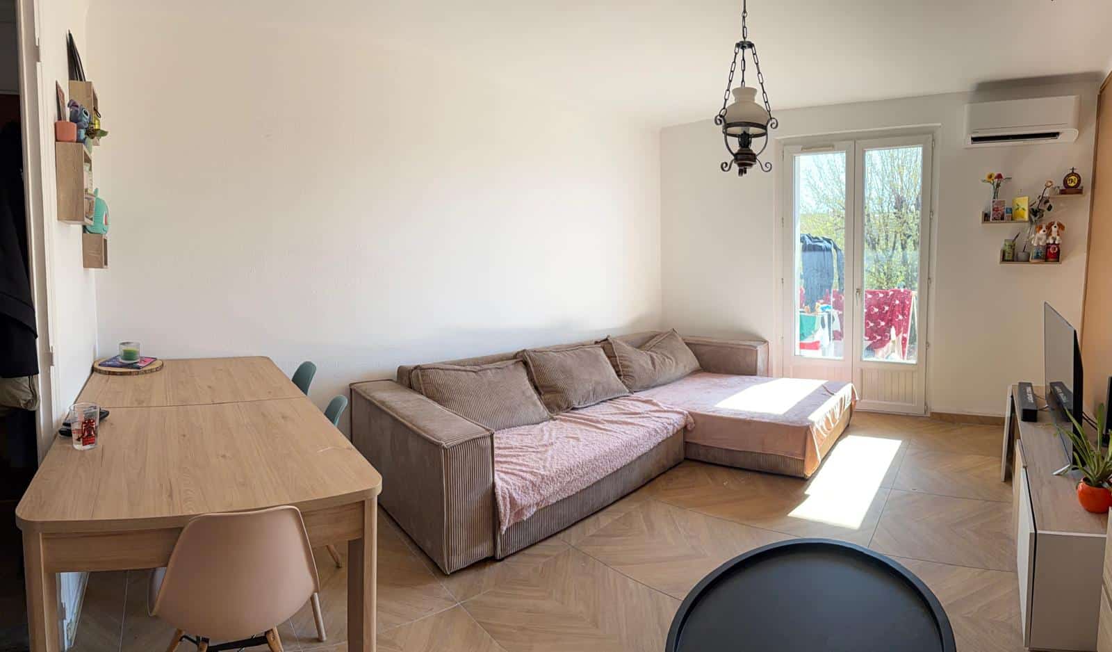 Cozy living room with a beige L-shaped sofa, a long wooden dining table, chairs, and a balcony door letting sunlight in.