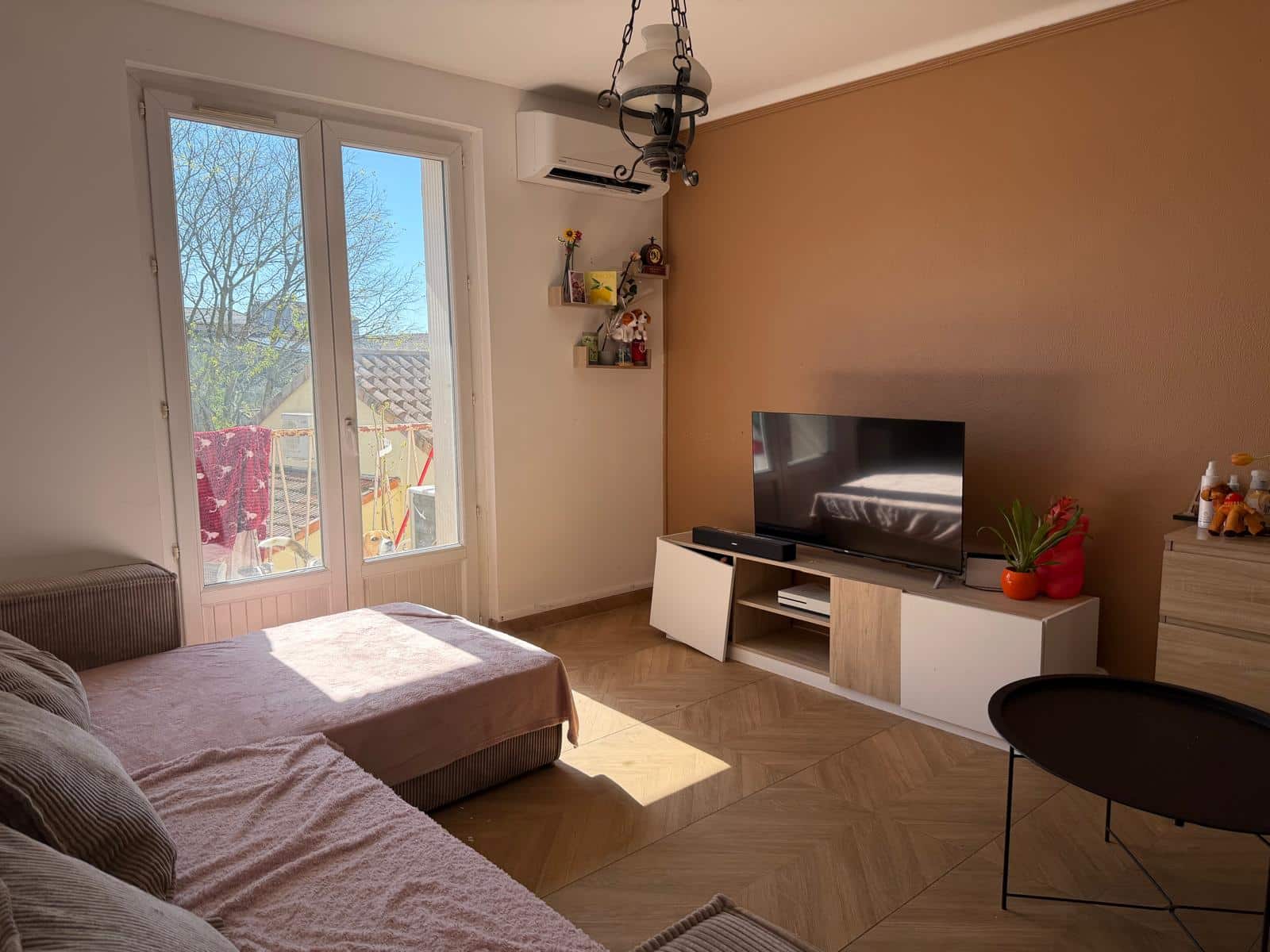 Living room with a wall-mounted TV, beige accent wall, and a glass door leading to a balcony bathed in sunlight. A small shelf with decorations hangs near an air conditioner above the door. A couch covered in a pink blanket sits in the foreground.