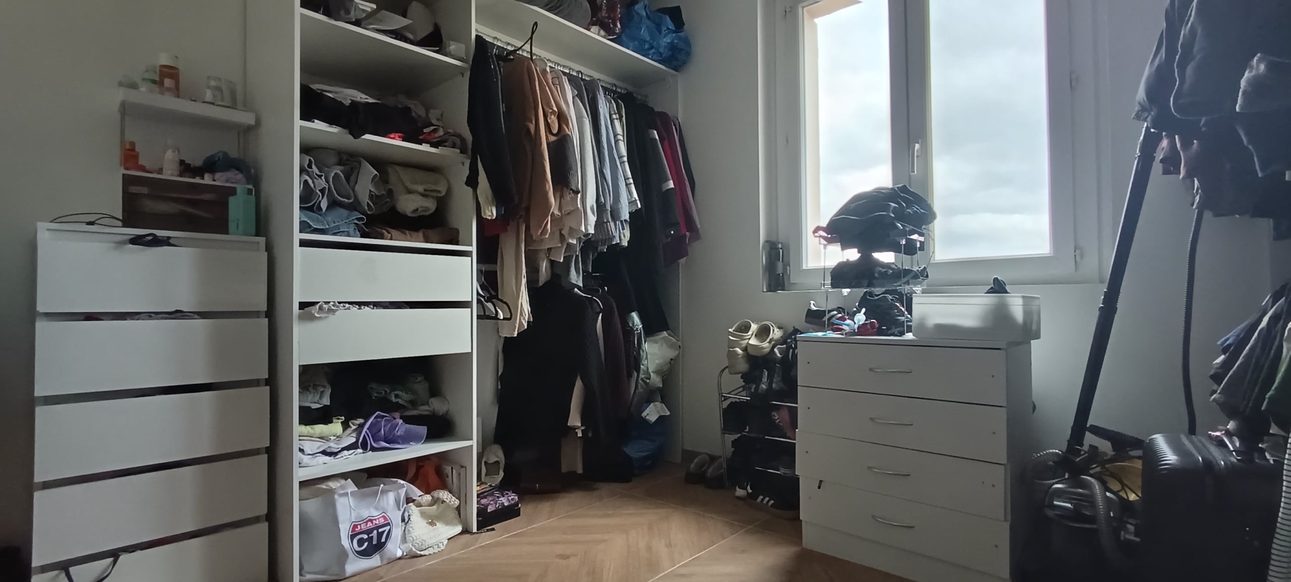 A cluttered walk‑in closet with shelves, hanging clothes, and a window on the right; drawers and shoe organizers below hold shoes and folded items.
